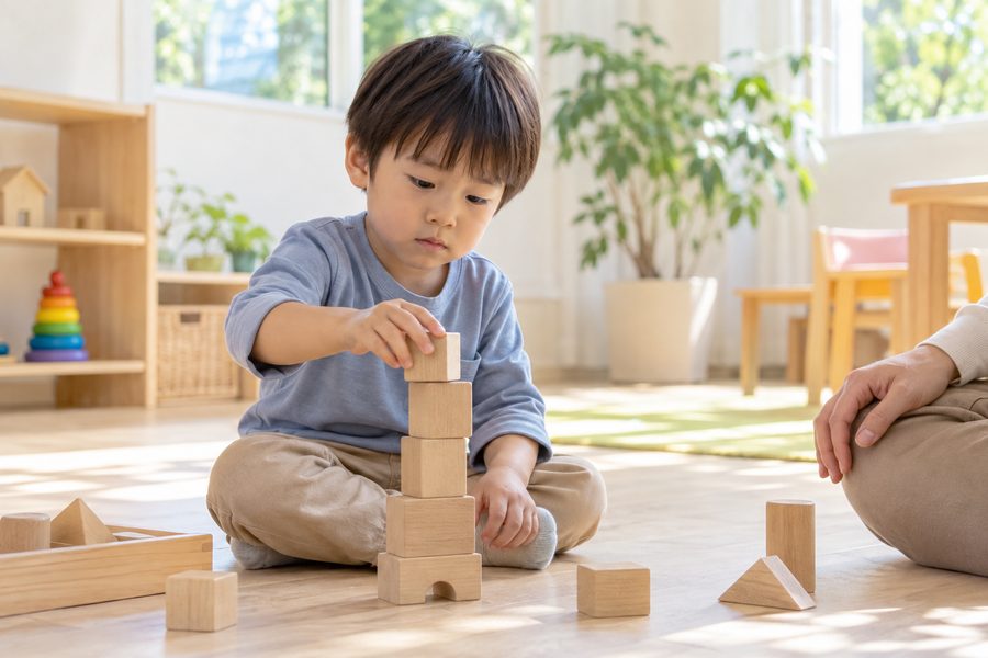 床に座って木の積み木を積む男の子と、そばで見守る大人の手